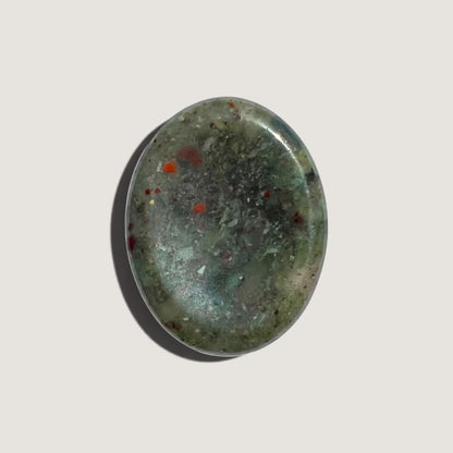 A polished oval bloodstone worry stone featuring a mottled pattern of dark green, light olive green and small red inclusions, displayed on a neutral light grey background.