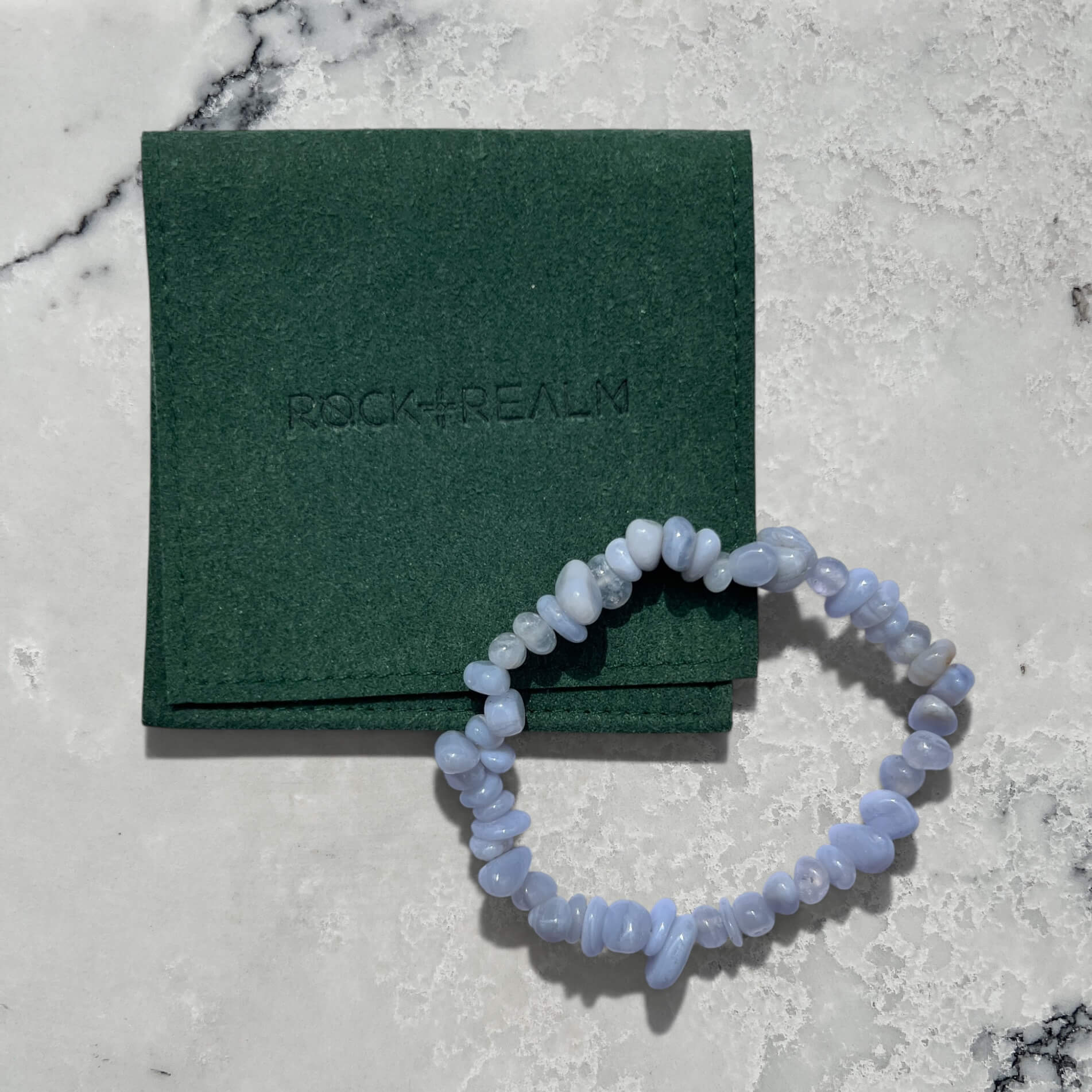 Blue Agate crystal chip gemstone bracelet on a marble surface with a green 'Rock + Realm' branded pouch