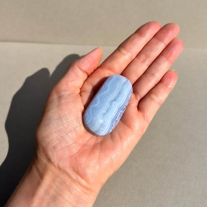An open hand holds a small oval blue lace agate palm stone displaying delicate agate banding in pale blue against a taupe backdrop