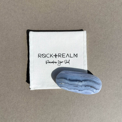 A polished oval blue lace agate palmstone with natural white banding, displayed on top of a white cotton branded 'ROCK + REALM Reawaken Your Soul' against a neutral taupe background.