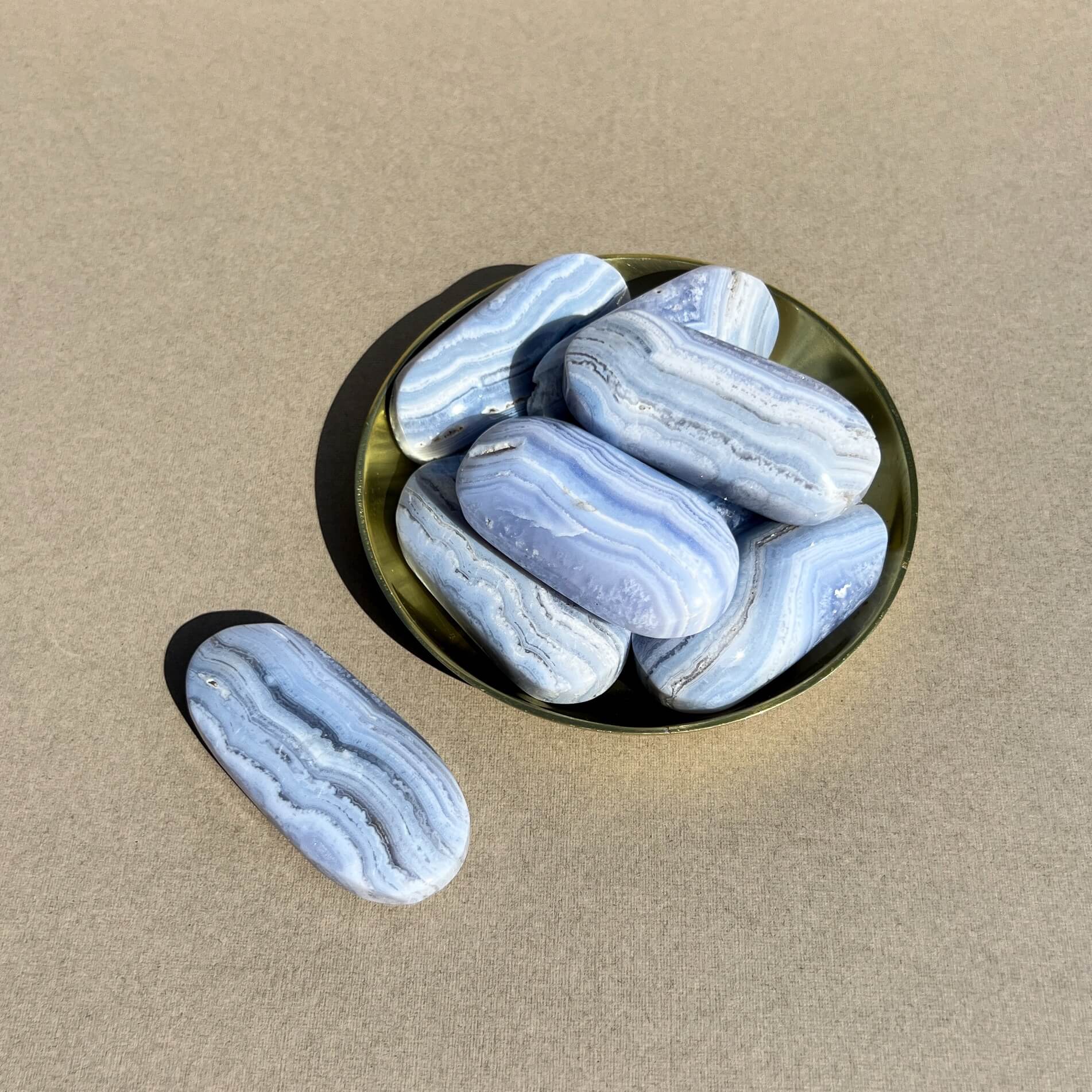 Several polished blue-and-white lace agate palmstones arranged in a small round brass dish, with one blue lace agate palmstone placed beside it on a beige surface.