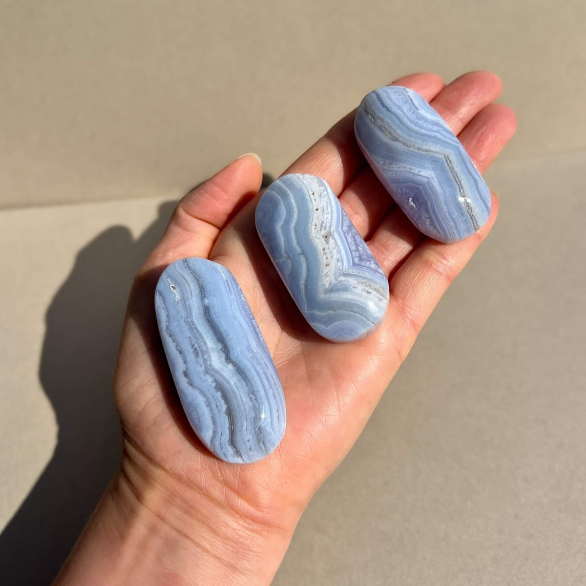 Three polished light blue lace agate palmstones with white veining are held in a person's hand against a neutral beige background.