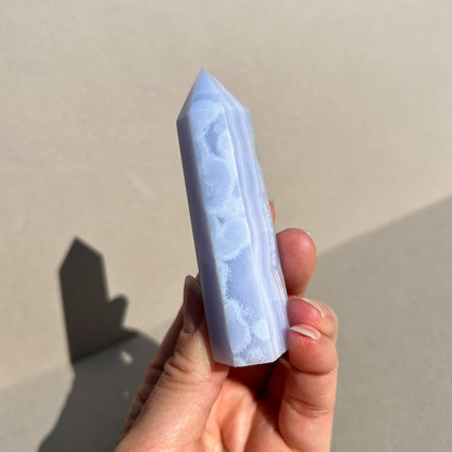 A hand holding a polished pale blue lace agate crystal point against a neutral beige background with a soft shadow.