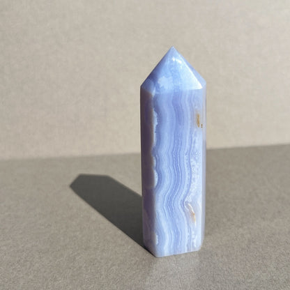 A polished blue lace agate crystal point tower standing upright on a neutral gray surface with a soft shadow cast to the left.