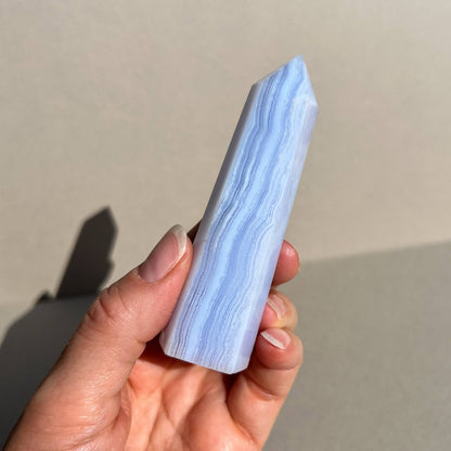 A hand holding a polished blue and white banded blue lace agate crystal tower against a neutral beige background.