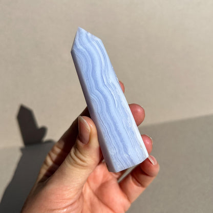 A hand holding a polished light blue and white blue lace agate crystal point against a neutral beige background.