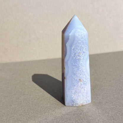 A polished natural blue lace agate crystal point with a light blue and white agate pattern, standing upright on a neutral gray surface with a soft shadow cast to the left.