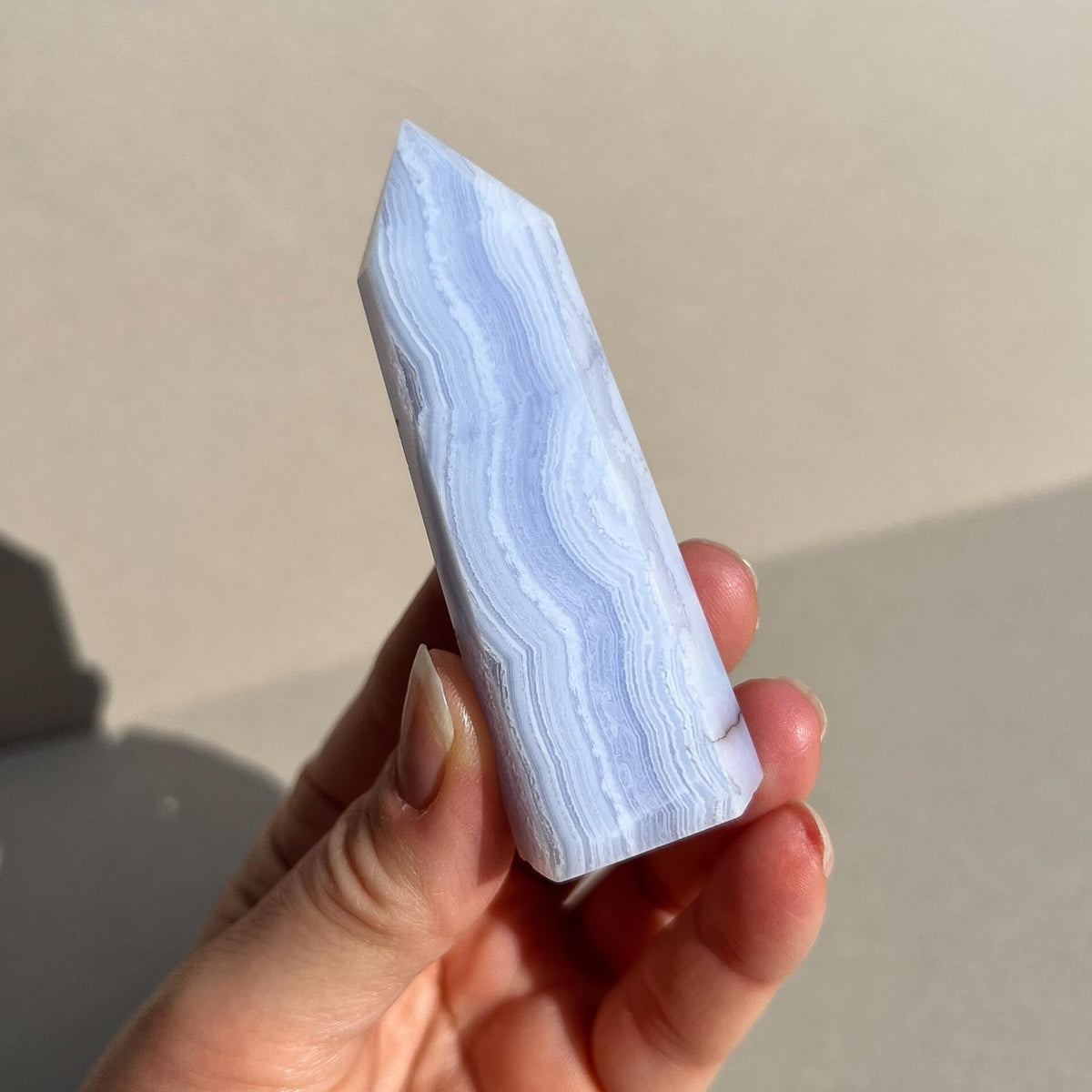 A hand holding a polished pointed crystal tower made of light blue and white banded blue lace agate, set against a neutral beige background with soft shadows.