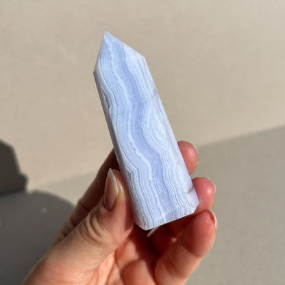 A hand holding a polished pointed crystal tower made of light blue and white banded blue lace agate, set against a neutral beige background with soft shadows.