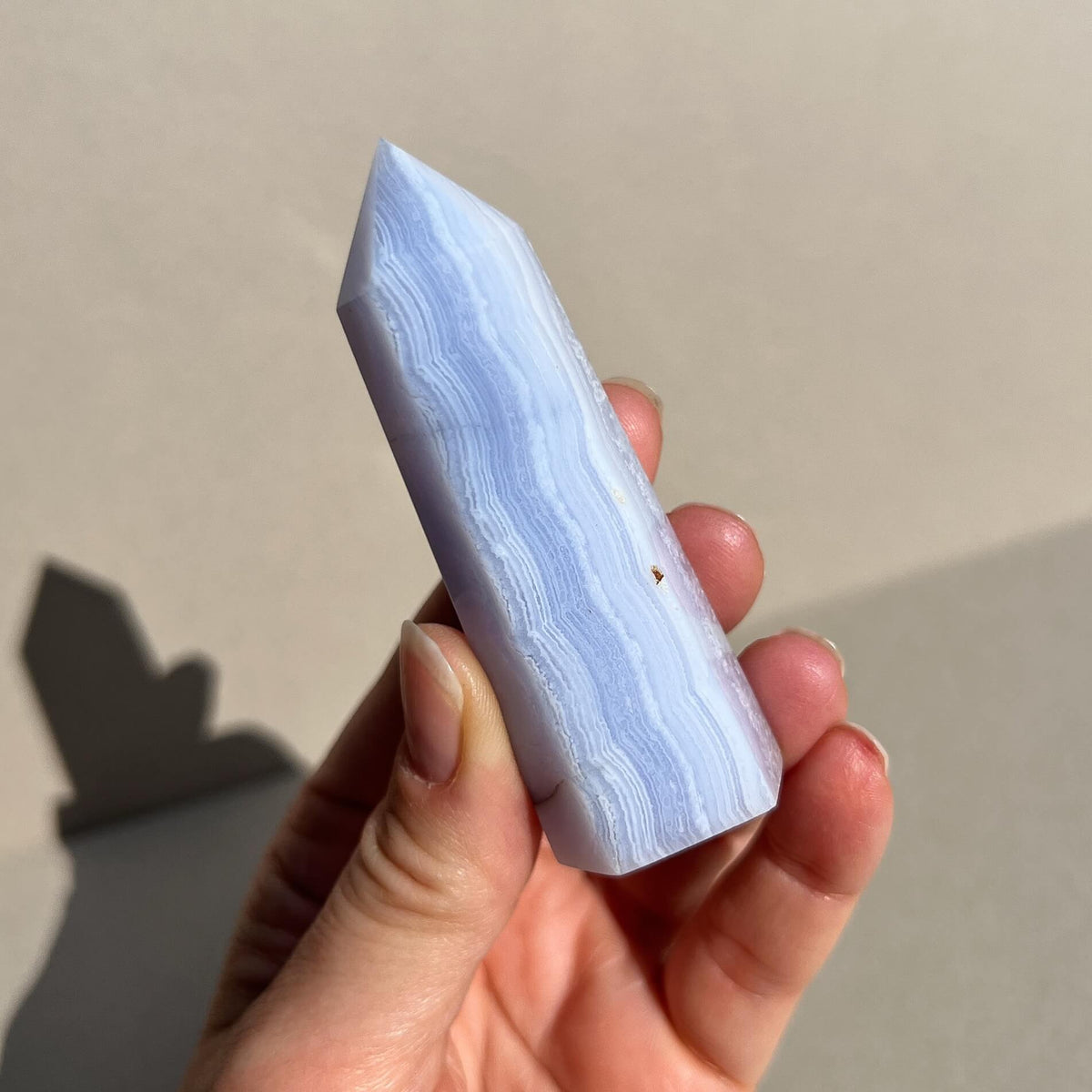 A hand holding a polished pointed blue lace agate crystal tower with light blue and white agate-like banding, photographed against a plain beige wall with a soft shadow.