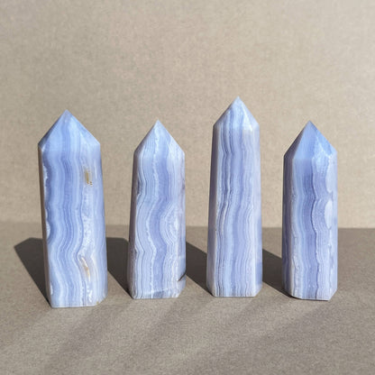 Four blue lace agate pointed crystal towers made of light blue and white marbled stone, arranged in a row against a neutral beige background.