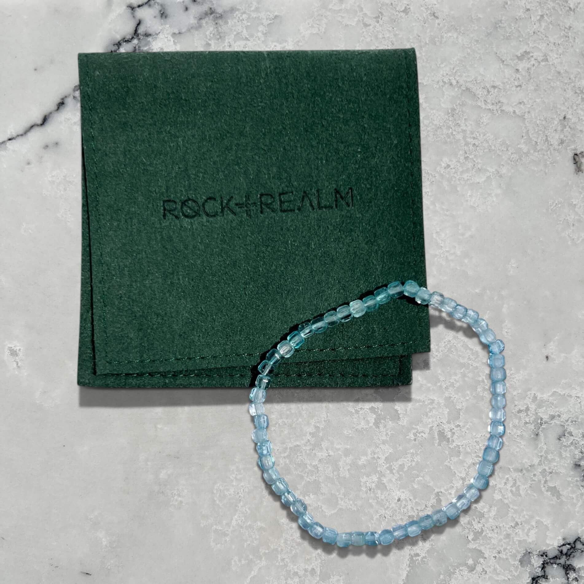 Blue Topaz gemstone bead bracelet on a green jewellery pouch with 'Rock + Realm' branding on a marble surface
