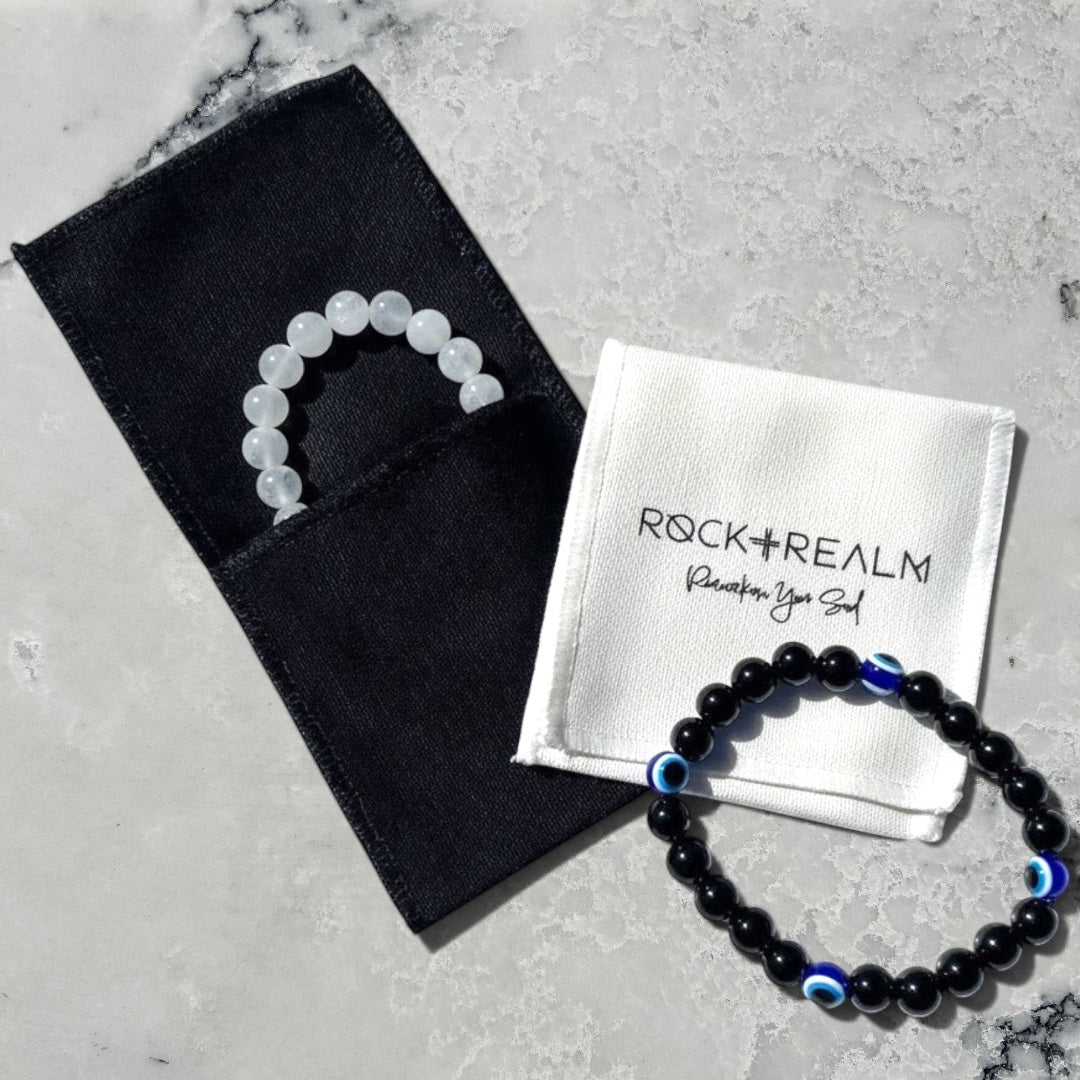 White selenite crystal bead bracelet in a black jewellery pouch overlay with a white 'Rock + Realm' branded cotton pouch with a black obsidian and blue evil eye beaded bracelet on a marble surface