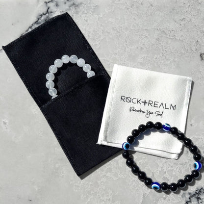 White selenite crystal bead bracelet in a black jewellery pouch overlay with a white 'Rock + Realm' branded cotton pouch with a black obsidian and blue evil eye beaded bracelet on a marble surface