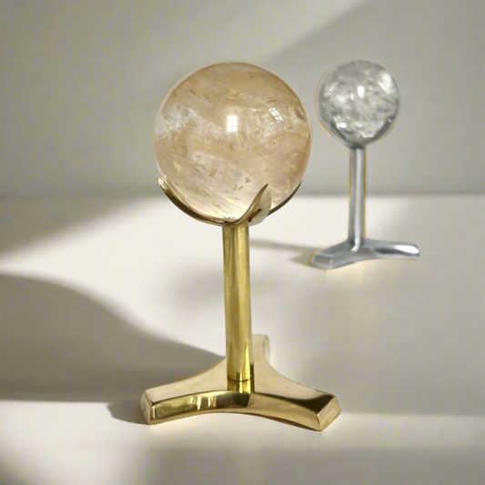 A large crystal sphere perched on an elegant tall brass display stand with a quartz crystal sphere on a silver display stand in the background