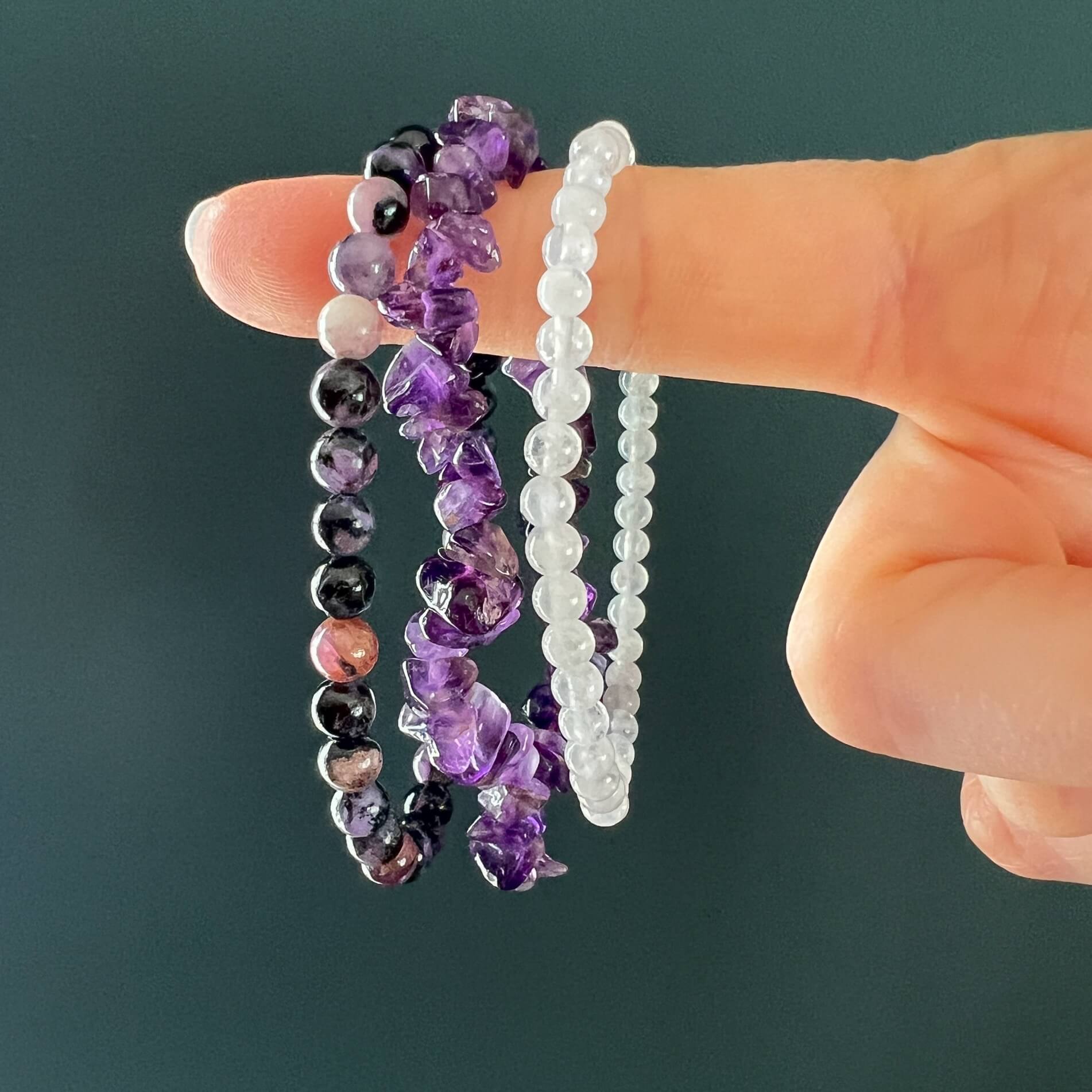 Hand holding three purple & white crystal bead bracelets against a dark green background