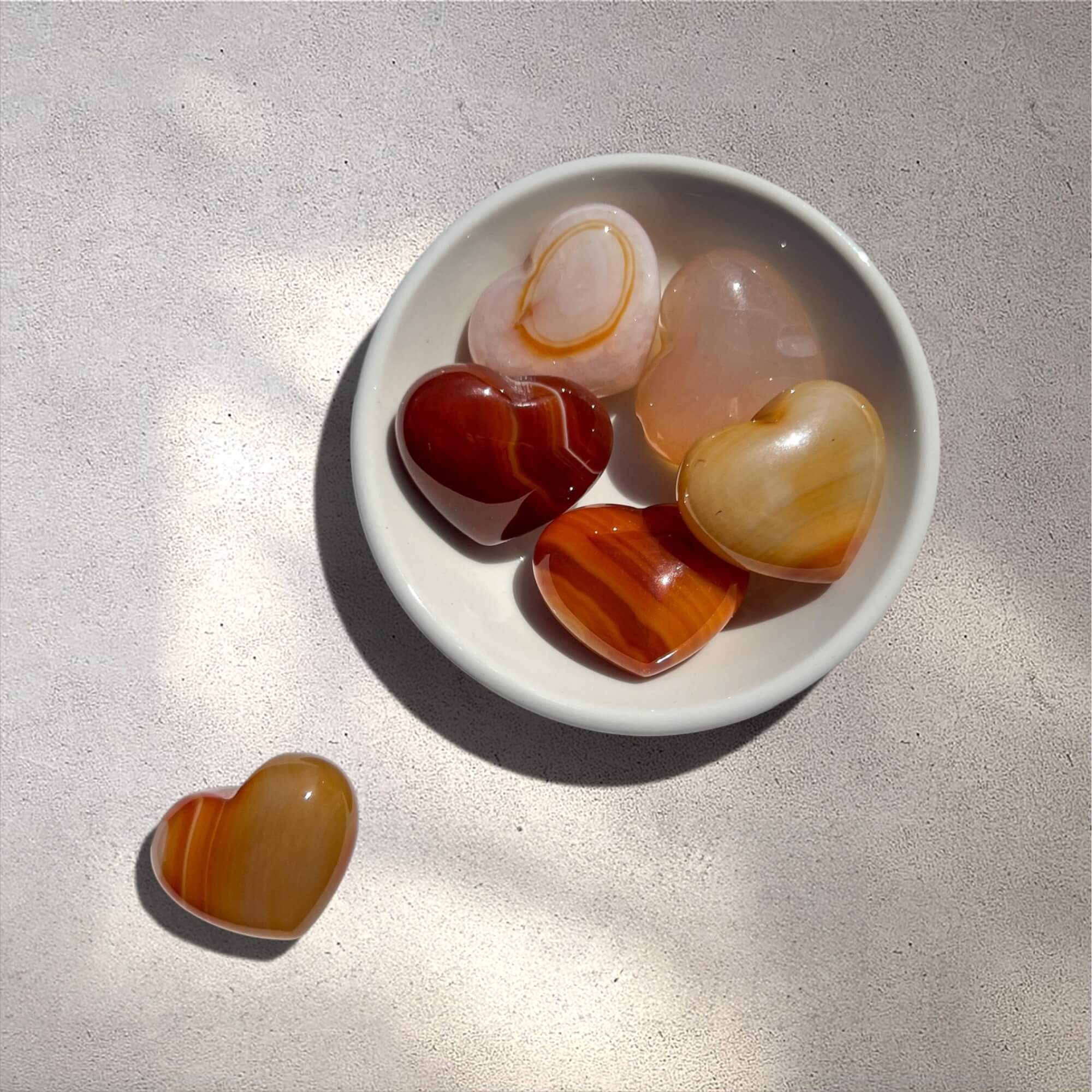 Five polished heart-shaped carnelian crystals in various colours, including amber, reddish-brown and pale pink, displayed in a small white ceramic bowl with one crystal placed outside the bowl on a textured light grey surface.