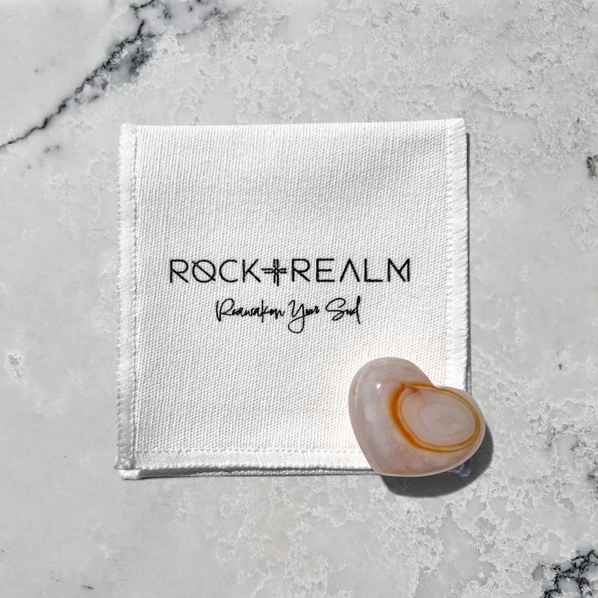 A polished heart-shaped carnelian crystal with orange and white banding, resting on a white cotton pouch printed with the brand name 'ROCK + REALM' and the tagline 'Reawaken Your Soul', all placed on a light grey marble surface.