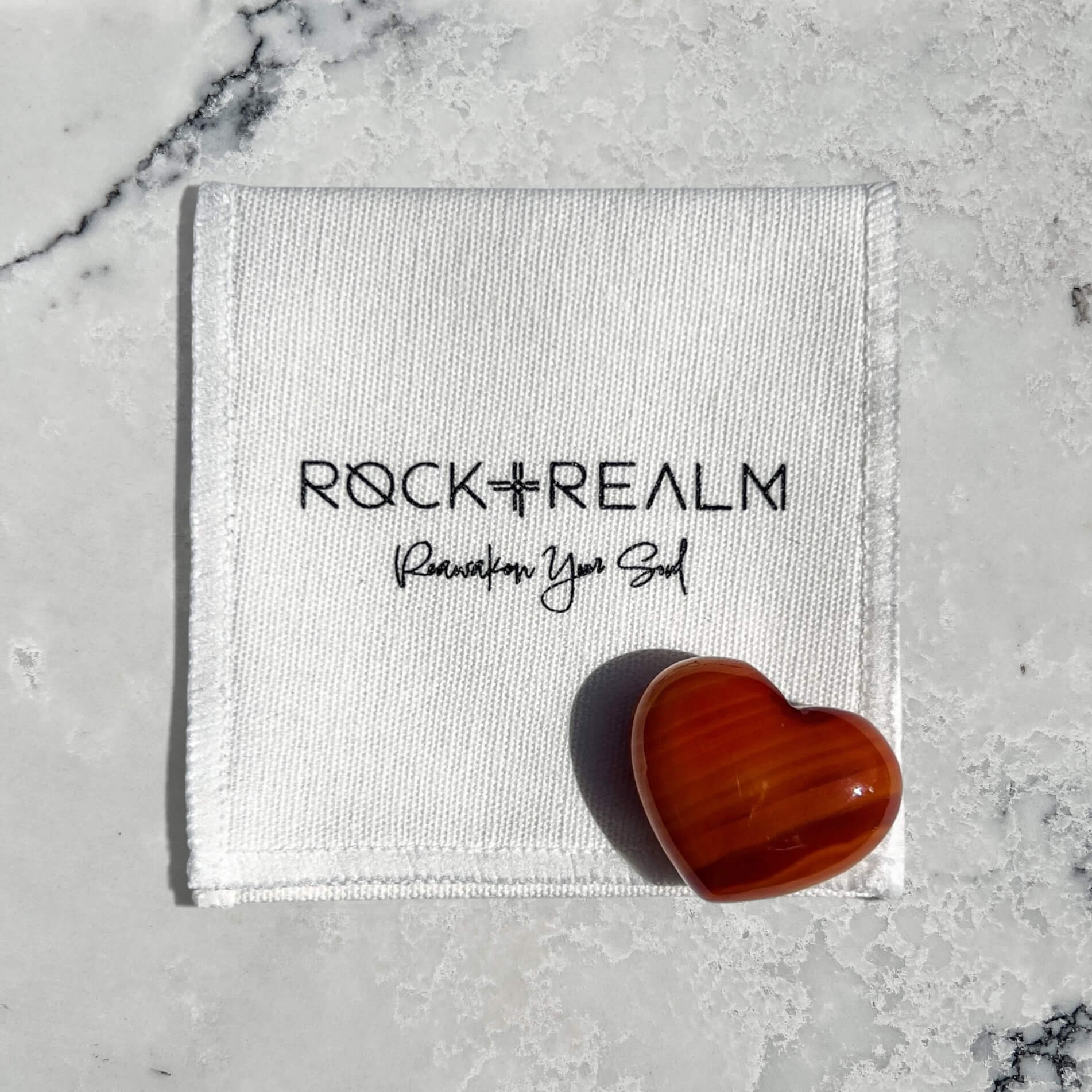A polished heart-shaped carnelian crystalrests on a white textured fabric pouch printed with the brand name 'ROCK + REALM' and the tagline 'Reawaken Your Soul', set against a light gray marble background.