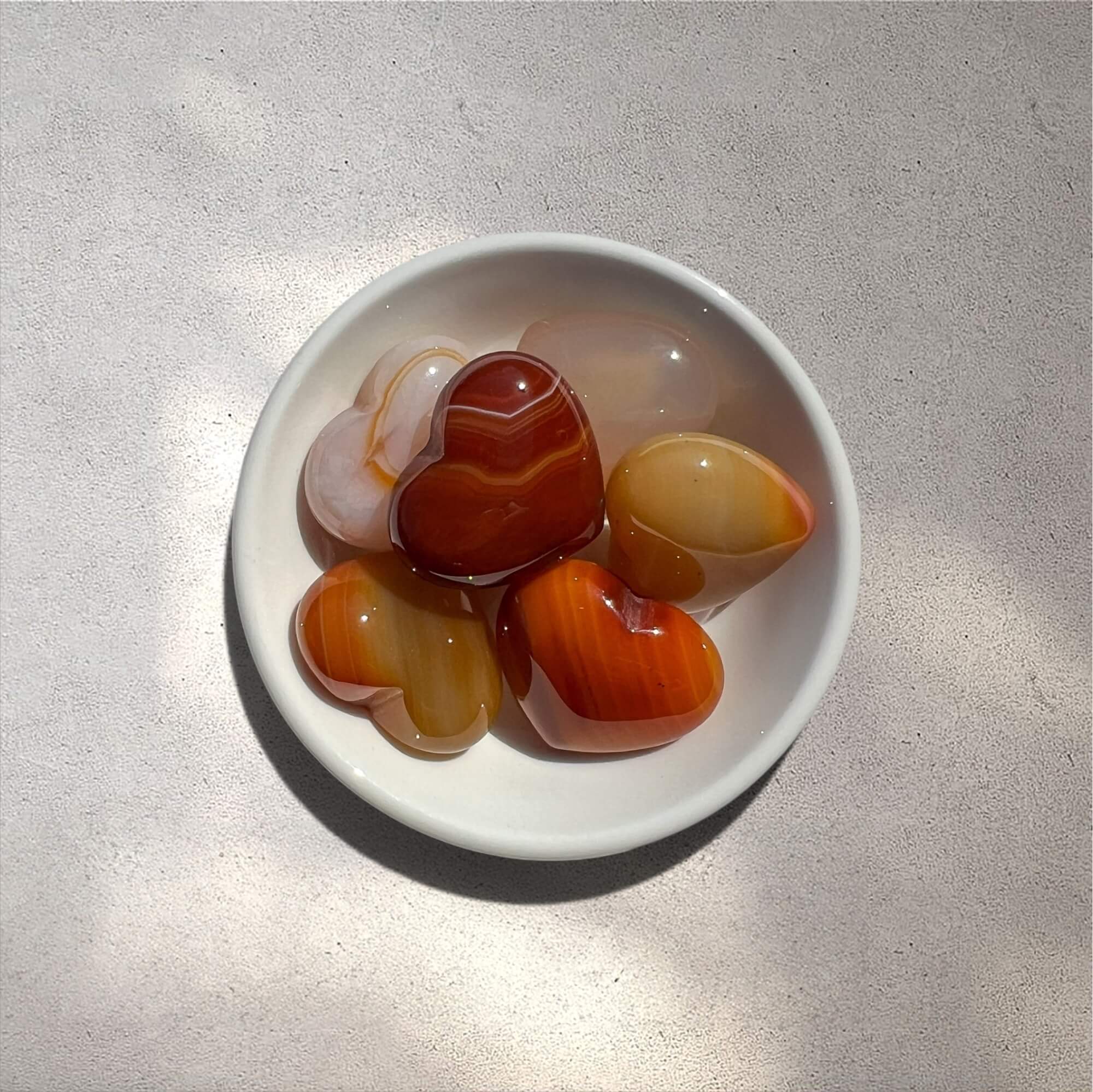 Five polished heart-shaped carnelian stones in various shades of orange, red and cream, arranged in a small white ceramic bowl on a textured light grey surface.