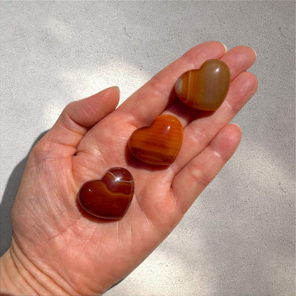 Three polished heart-shaped carnelian crystals with natural red and orange banding are displayed on an open hand against a textured grey concrete background.