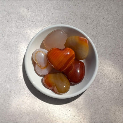 A white ceramic bowl filled with polished heart-shaped carnelian stones in various warm tones of orange, red, brown and cream, set on a textured light beige surface with soft natural shadows.