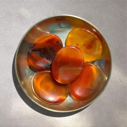 Five polished carnelian oval worry stones with swirling orange, red and brown banding, arranged inside a round brass dish on a textured grey surface.