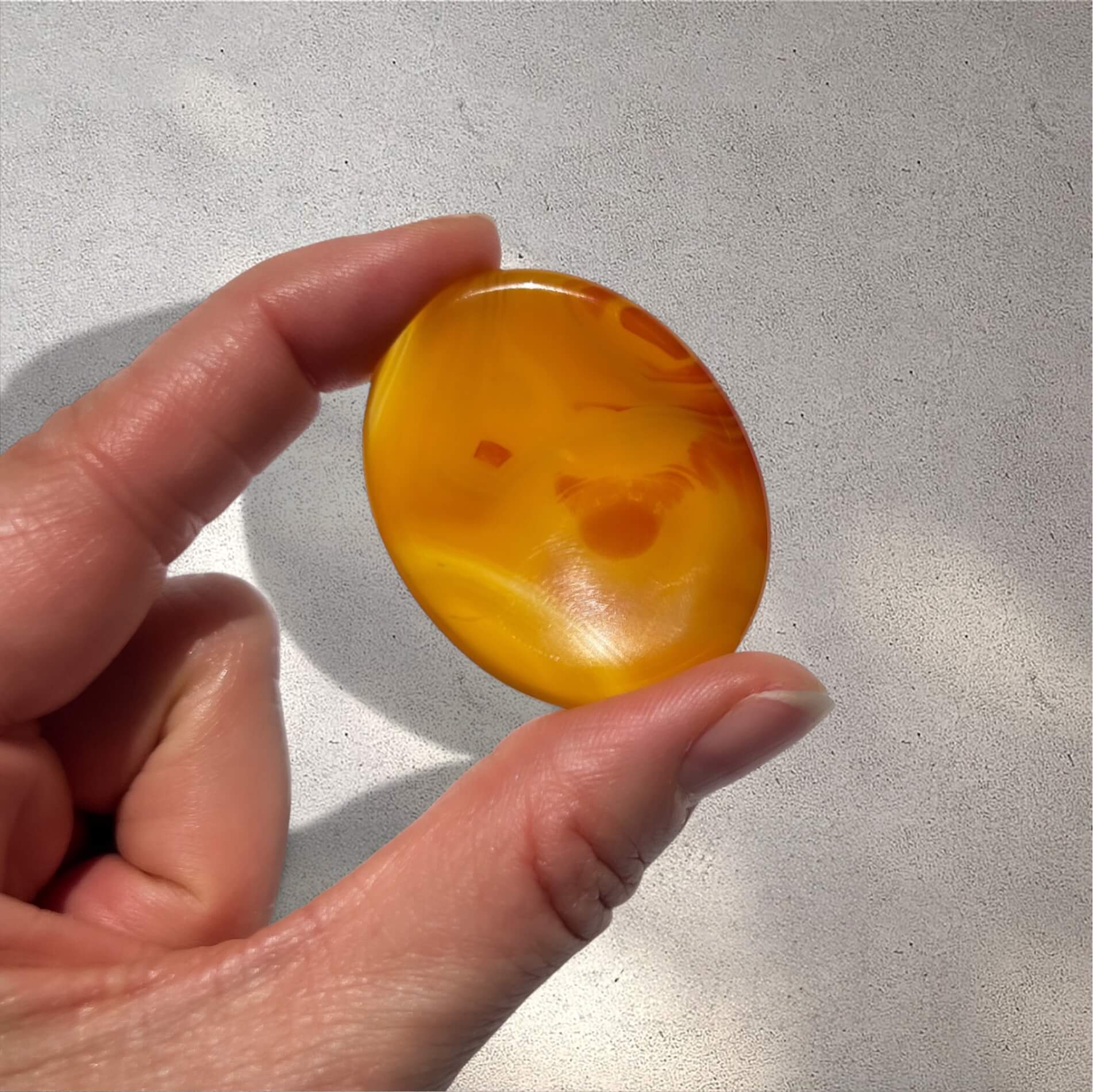 A hand holding a smooth, polished oval orange carnelian worry stone against a light grey textured background.