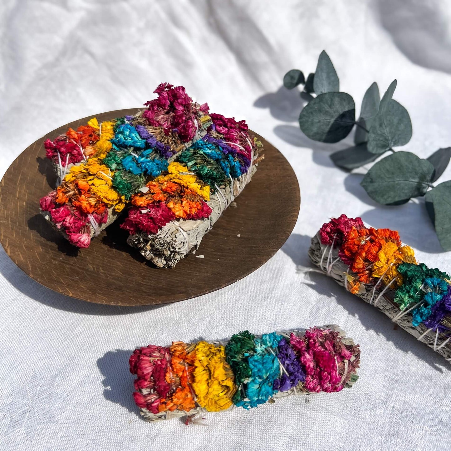 Bright rainbow coloured 7 chakra sage smudge bundles in a brown wooden dish on a white cloth with eucalyptus in the background