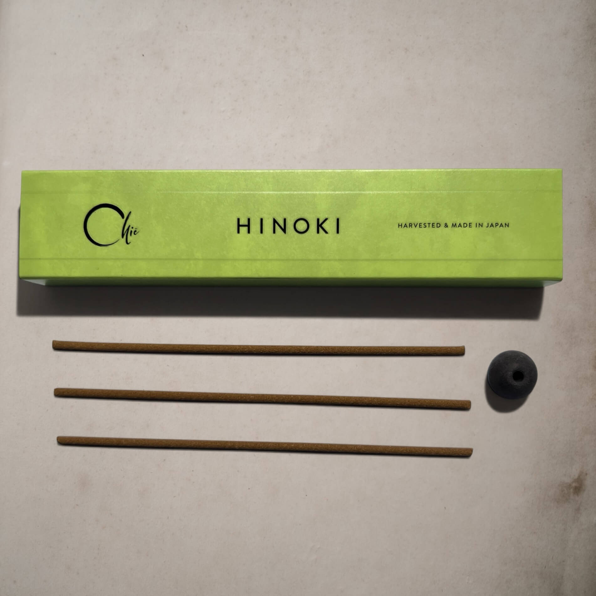 A lime green rectangular box labeled 'Chie HINOKI' with three brown incense sticks and a small black incense holder placed below on a light beige surface.