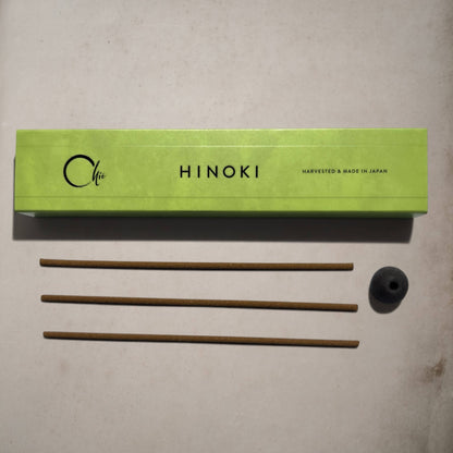 A lime green rectangular box labeled 'Chie HINOKI' with three brown incense sticks and a small black incense holder placed below on a light beige surface.