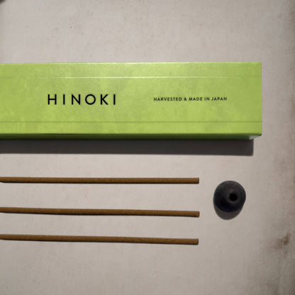 A Hinoki incense set on a light beige surface, featuring a lime green box with black lettering, four brown incense sticks, and a small black incense holder.