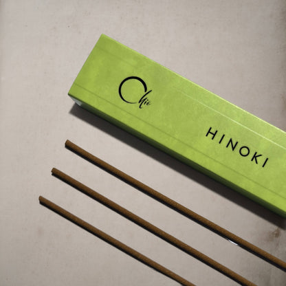 A bright lime green rectangular box labeled 'Chie HINOKI' lies diagonally on a light beige surface, with three thin brown incense sticks placed below it.