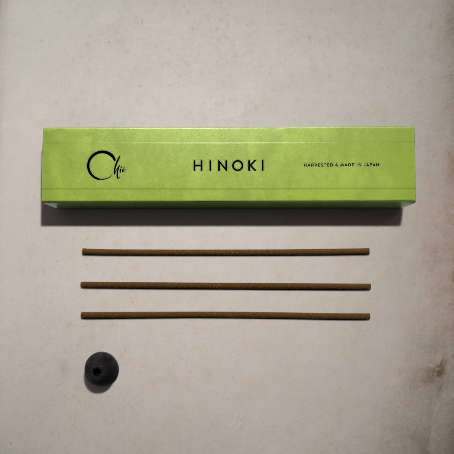 A light green rectangular box labeled 'Hinoki' with 'Harvested & Made in Japan' printed on it, placed on a light beige surface above three thin brown incense sticks and a small black round incense holder.
