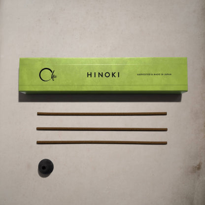 A light green rectangular box labeled 'Hinoki' with 'Harvested & Made in Japan' printed on it, placed on a light beige surface above three thin brown incense sticks and a small black round incense holder.