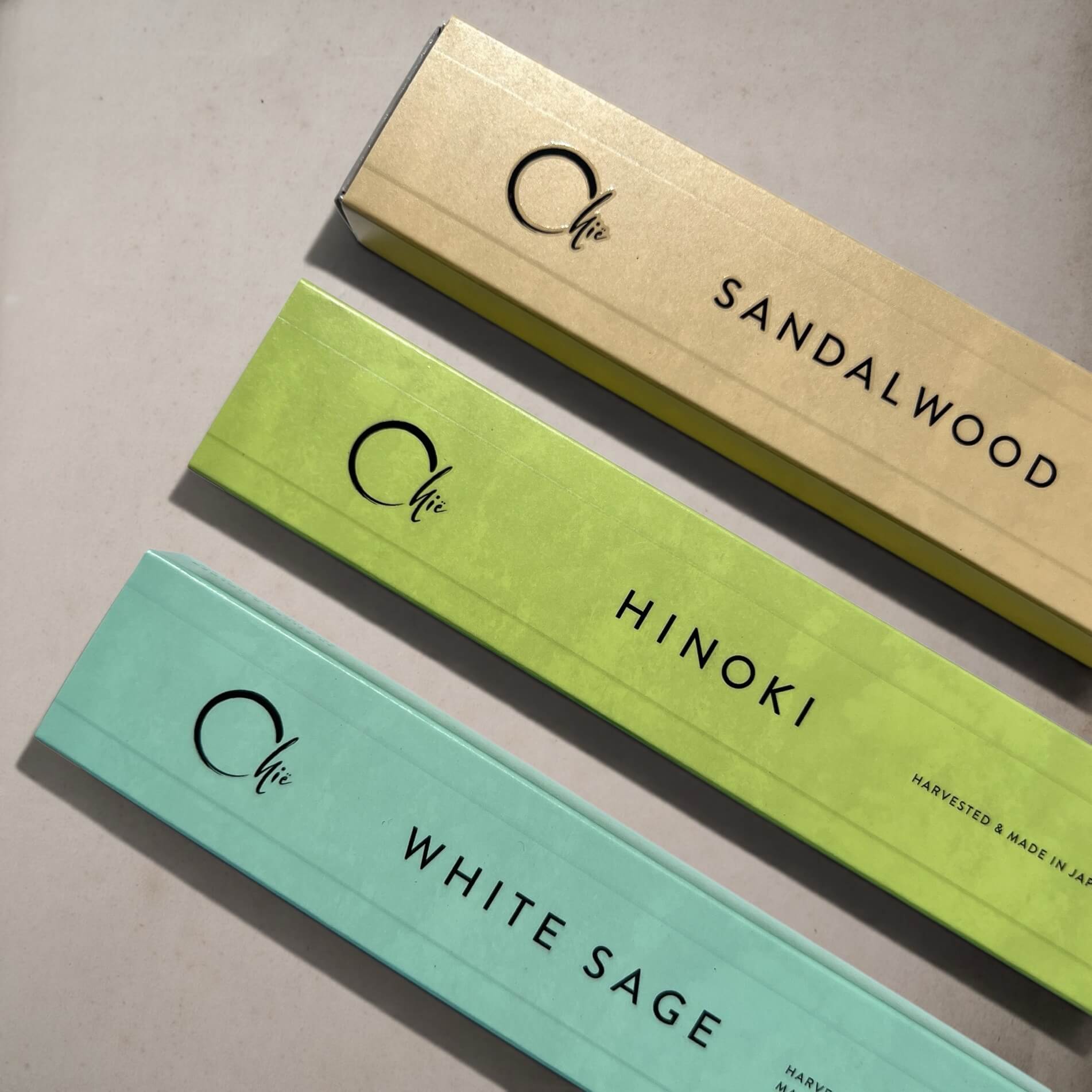 Three long rectangular product boxes arranged diagonally on a light neutral surface. The boxes are labeled with the brand name 'Chie' and scent names 'SANDALWOOD', 'HINOKI', and 'WHITE SAGE', with colours corresponding to sandalwood, hinoki and white sage.