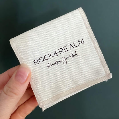 'Rock + Realm' branded white cotton jewellery pouch held against a dark green background