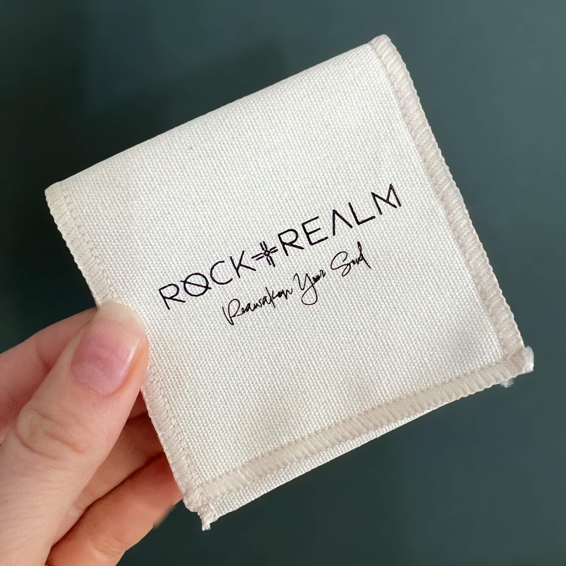 'Rock + Realm - Reawaken Your Soul' Branded white cotton pouch held in front of a teal backdrop