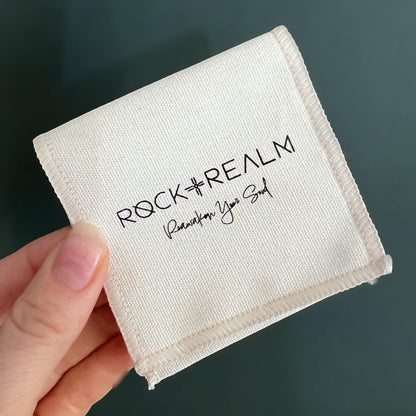 White cotton square jewellery pouch with 'Rock + Realm' branding held by a hand against a dark background