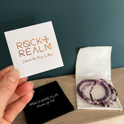 'Rock + Realm' copper foil branded lux card held to camera with two branded cotton jewellery pouches and a stack of pink and purple bead bracelets in the background with a teal backdrop