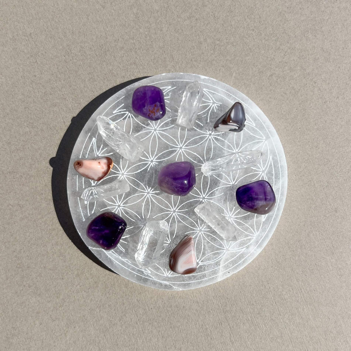 A circular white selenite charging plate with etched geometric patterns, holding several polished tumbled stones including purple amethysts, Botswana agate and clear quartz in a crystal grid formation, photographed on a neutral beige surface.