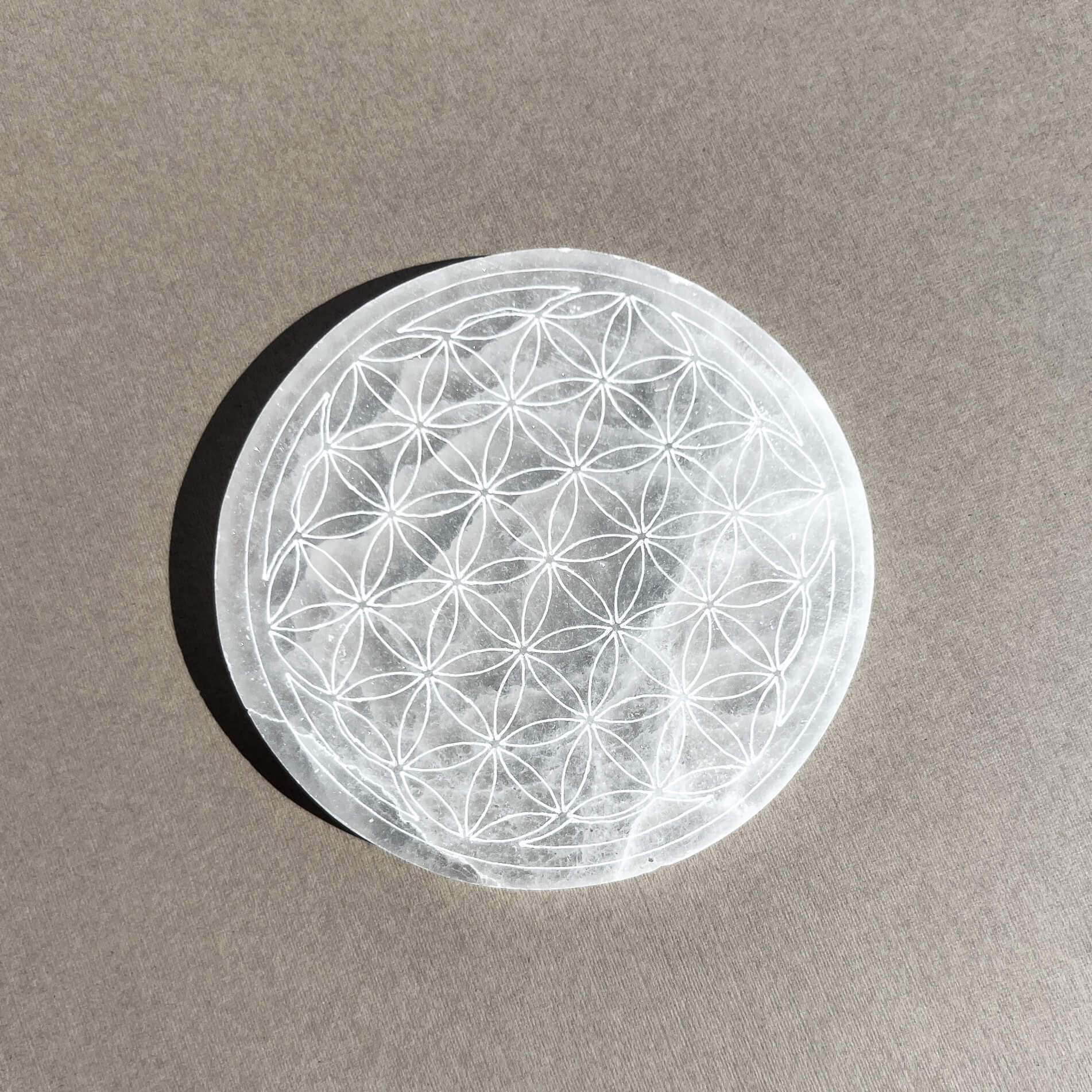 A circular selenite plate featuring an intricate geometric flower of life pattern, resting on a textured grey surface with a soft shadow.