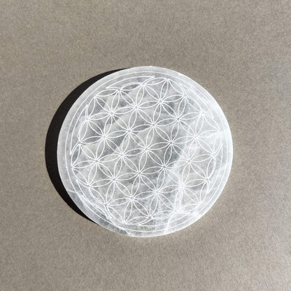 A circular selenite plate featuring an intricate geometric flower of life pattern, resting on a textured grey surface with a soft shadow.