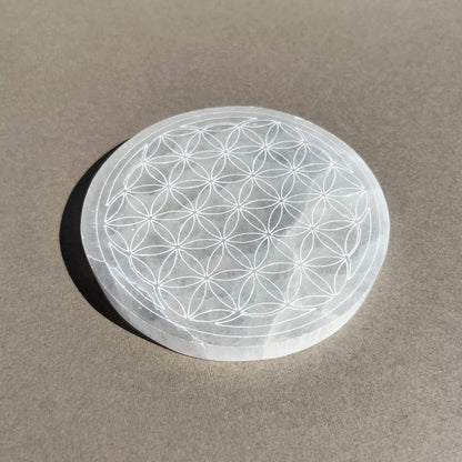 A circular selenite plate featuring an etched Flower of Life geometric pattern, resting on a textured grey surface.