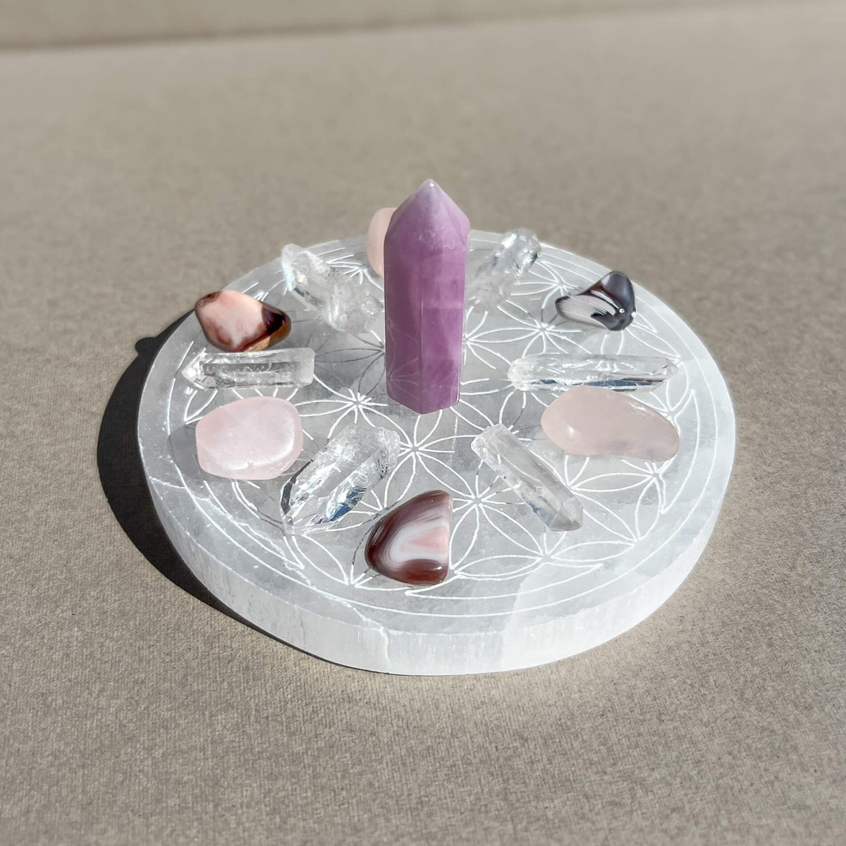 A crystal grid set arranged on a white circular selenite charging plate with an engraved geometric flower pattern. The set includes a mini kunzite tower, several clear quartz points, pink Botswana agate and rose quartz tumbles, photographed on a neutral beige surface in bright sunlight.