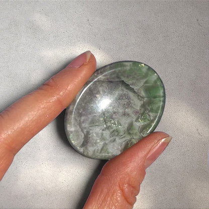 Polished fluorite worry stone with green and purple swirls, featuring a thumb indentation for stress relief held between thumb and forefinger