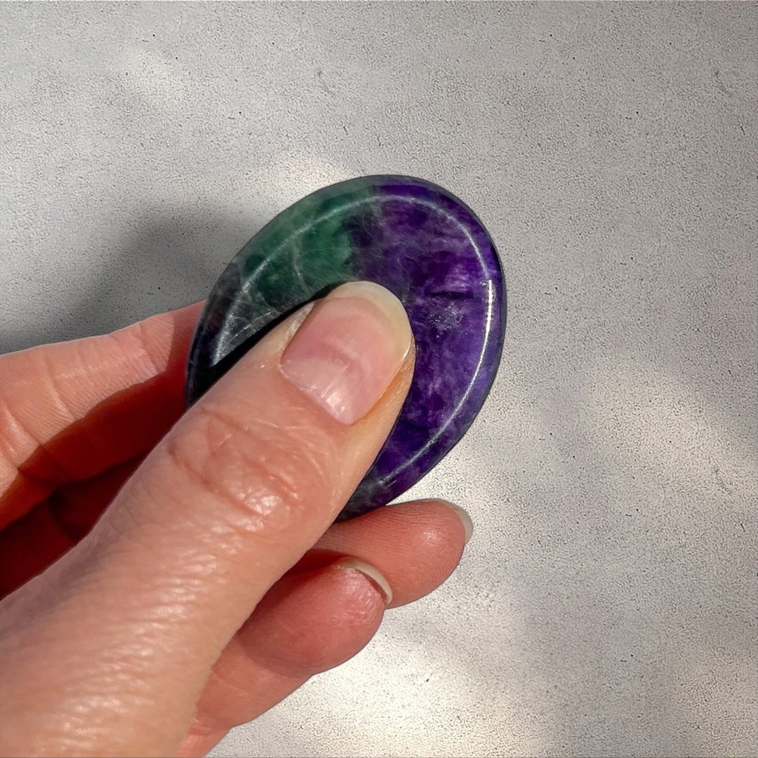 Polished fluorite worry stone with green and purple bands, held by a thumb and forefinger