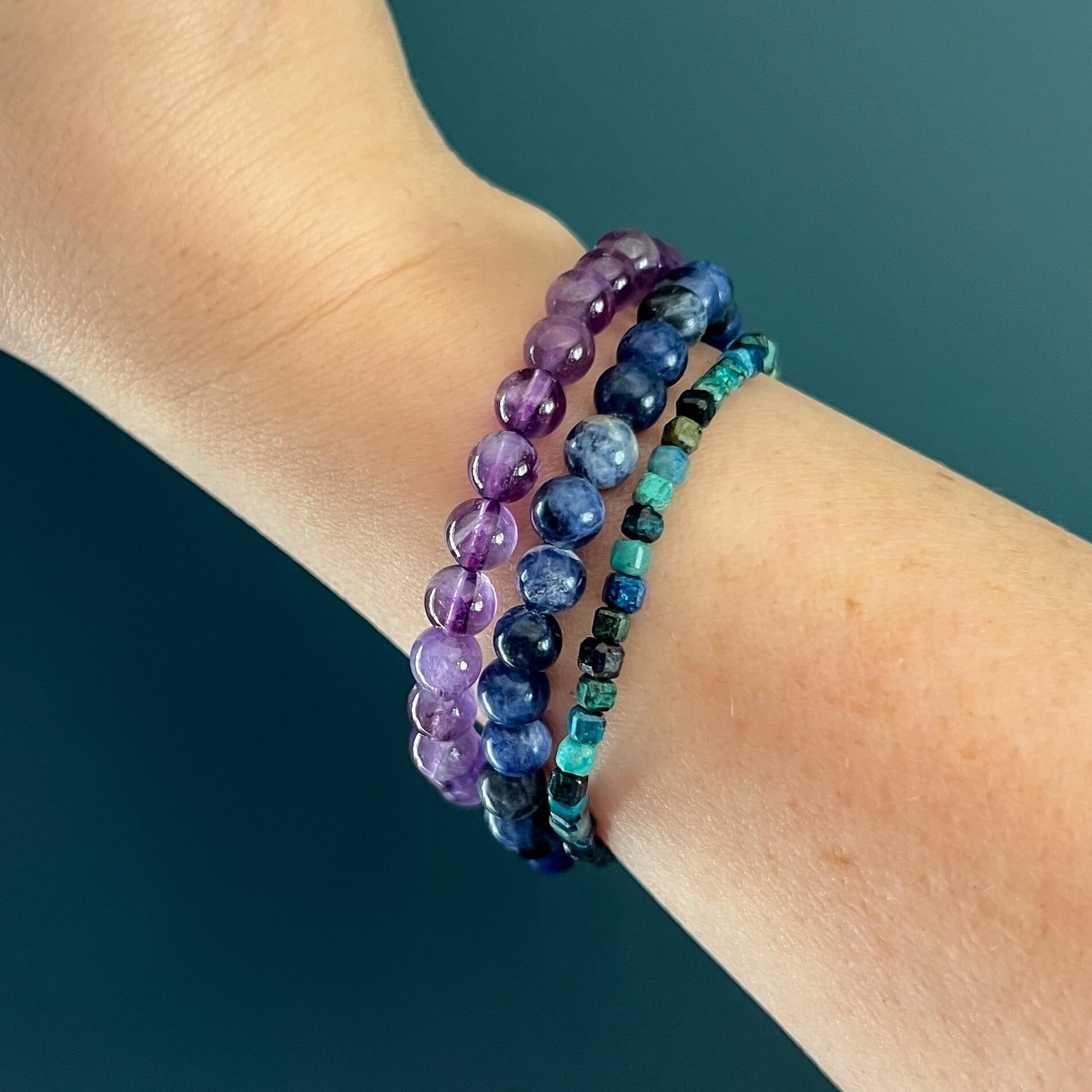 Three crystal bead bracelets in purple, blue and green on a wrist against a teal background
