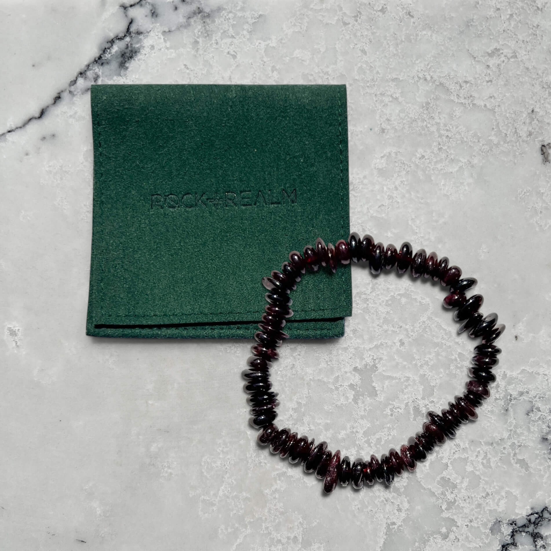 Garnet crystal beaded bracelet on a gray surface with a green pouch labeled 'Rock + Realm'.