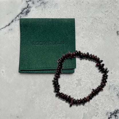 Garnet crystal beaded bracelet on a gray surface with a green pouch labeled 'Rock + Realm'.